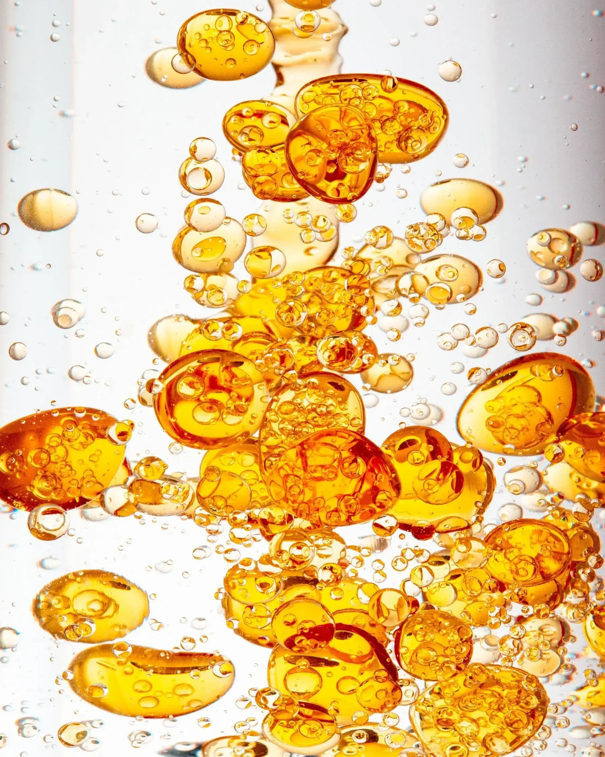 Close-up of golden-orange oil droplets of different sizes, with tiny bubbles suspended in a clear liquid against a white background