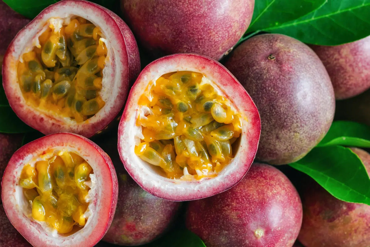 Close-up of whole and cut-open passion fruits with reddish-purple skins, golden-yellow juicy flesh and small dark seeds, surrounded by green leaves.