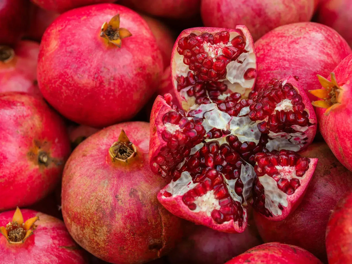 Fresh whole red pomegranates surrounding a cut-open pomegranate with juicy ruby-red seeds and white inner membranes